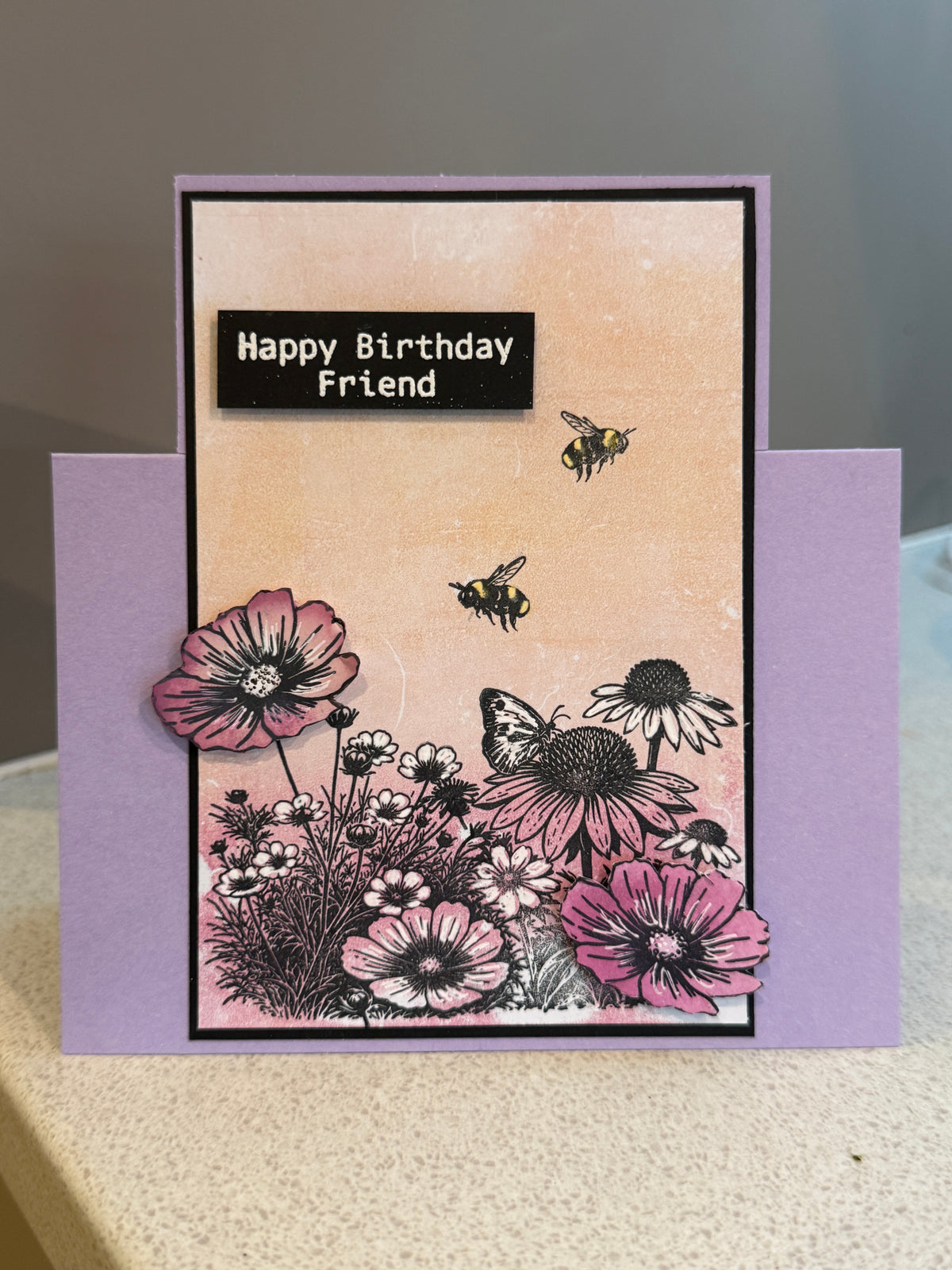 A handmade square birthday card with a lavender envelope, featuring a peach background, black and pink flowers, bumblebees, and a butterfly with the text 'Happy Birthday Friend'.