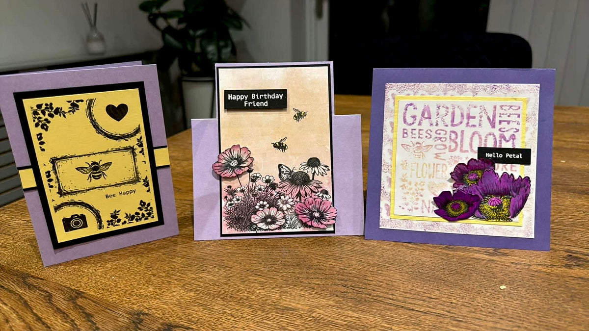 Three handmade greeting cards displayed on a wooden table. The left card has a lavender border and yellow design with black text and bee motifs.